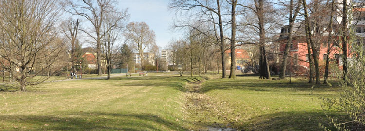 Blick vom Sportplatz in den Brunschwigpark, der vom Lindengraben durchzogen wird. Hier ist deutlich die Lage erkennbar: Die Bauhaus-Villa liegt bereits im Brunschwig-Park. Die Lessingstraße biegt noch vor dem Grundstück Richtung Stadthalle ab, nur der Radweg führt geradeaus weiter. Die beiden sich kreuzenden Radwege (links) gehören zu den meist befahrenen Radwegen in der Stadt, an der Kreuzung ist höchste Vorsicht geboten